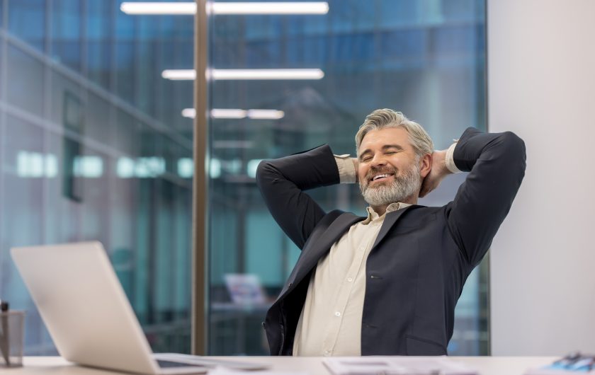 Mature businessman smiling, leaning back in his office chair with hands behind head, experiencing satisfaction and a brief break from work in a modern corporate environment