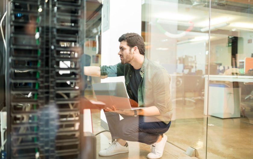Network administrator with notebook computer sitting in data center room and working with networking device on rack cabinet