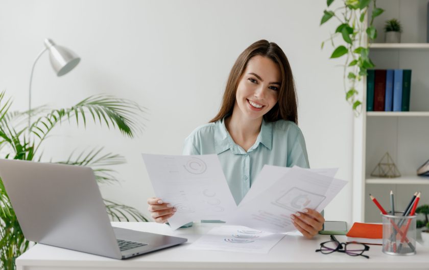 Smiling happy young successful employee business woman in blue shirt hold paper account documents sit work at workplace white desk with laptop pc computer at office indoors. Achievement career concept