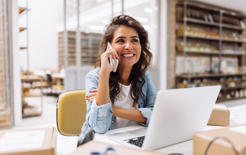 Cheerful businesswoman speaking on the phone while working in a warehouse. Happy online store owner making plans for product shipping. Female entrepreneur running an e-commerce small business.